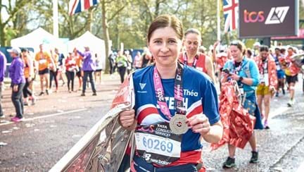 Stephanie wearing a Blind Veterans UK T-shirt with her name on posing for a photo after completing the marathon. In one hand she is holding a foil blanket and around her neck is her 2023 London Marathon medal