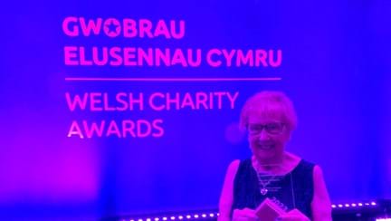Gill stands in front of a branded screen and holds her award proudly while smiling