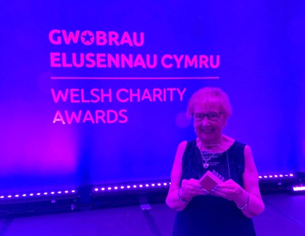 Gill stands in front of a branded screen and holds her award proudly while smiling
