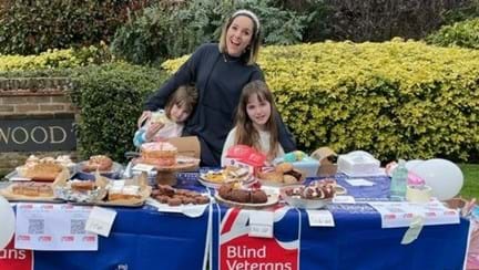 Mum Rachel with her arms around Mollie and Poppy as they stand behind a table full of cakes and Blind Veterans UK flags 