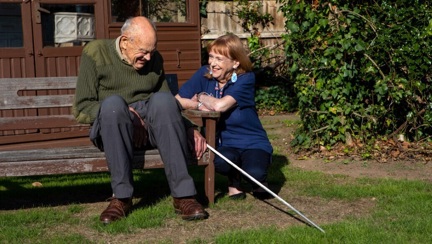 Mike sits on a bench and Jennie crouches next to him on a beautiful sunny day