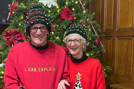 A man and woman wearing Christms jumpers and Christmas tree hats stand in front of a large Christmas tree