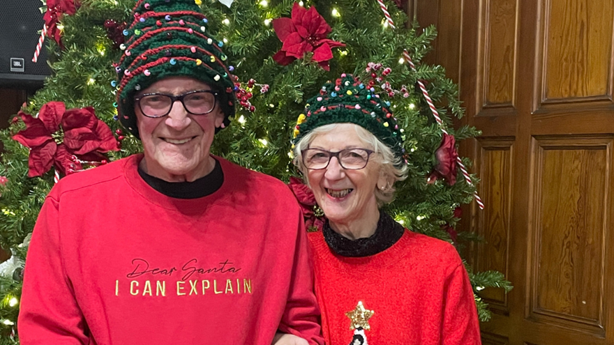 A man and woman wearing Christms jumpers and Christmas tree hats stand in front of a large Christmas tree