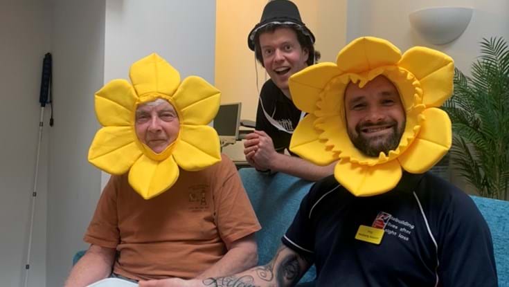 Two men sit on a sofa wearing daffodil costumes and a man stands behind them wearing a Welsh bonnet