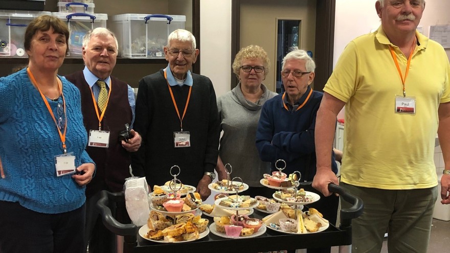 A group of blind veterans smiling, as they stand with a trolley of food they prepared 