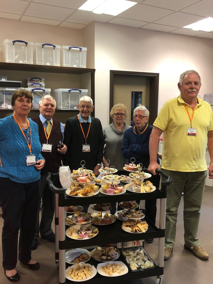 A group of blind veterans smiling, as they stand with a trolley of food they prepared