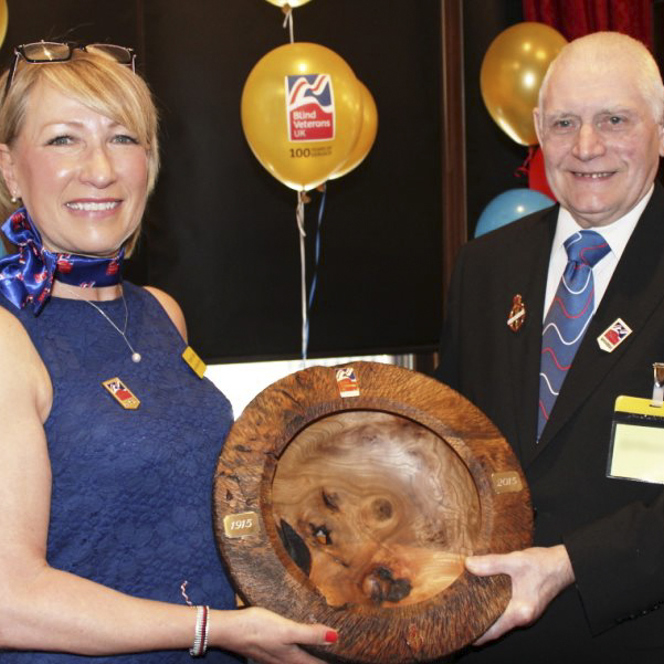 Blind veteran Bill and social worker Elizabeth smiling as they hold a wooden carved bowl