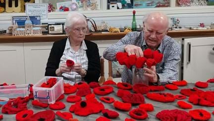 George and Shirley are sat at a table full of knitted poppies in the craft room at the Llandudno Centre