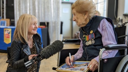 Margaret is wearing her medals and speaking into a microphone held by a journalist from the BBC