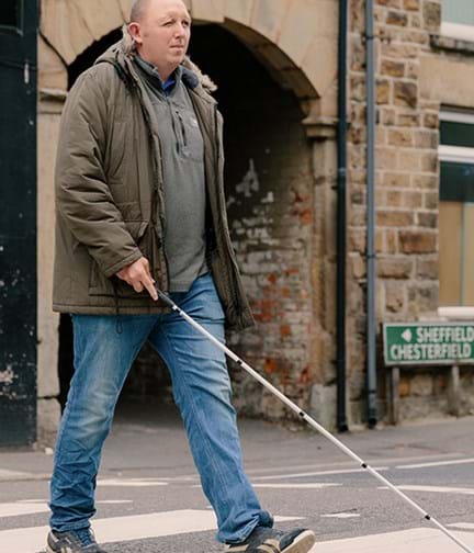 Blind veteran Chris crossing a the road at a zebra crossing using a white cane.