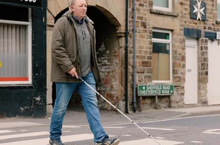 Blind veteran Chris crossing a the road at a zebra crossing using a white cane.
