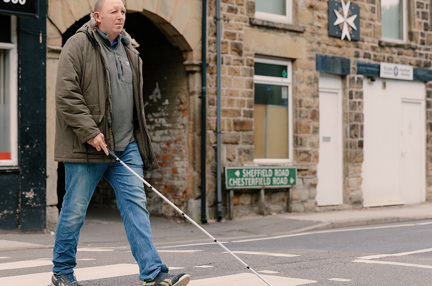 Blind veteran Chris crossing a the road at a zebra crossing using a white cane.