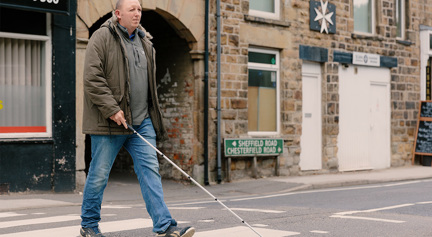 Blind veteran Chris crossing a the road at a zebra crossing using a white cane.