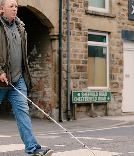 Blind veteran Chris crossing a the road at a zebra crossing using a white cane.