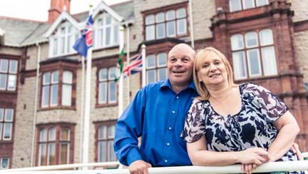 Blind veteran Billy with his arm around his wife, smiling as they stand outside our Centre of wellbeing in Llandudno