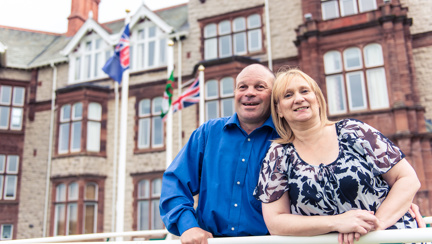 Blind veteran Billy with his arm around his wife, smiling as they stand outside our Centre of wellbeing in Llandudno