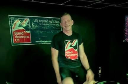 Ian sat on his spin bike wearing his Blind Veterans UK t shirt and with the Blind Veterans UK banner on the wall behind.