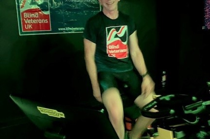 Ian sat on his spin bike wearing his Blind Veterans UK t shirt and with the Blind Veterans UK banner on the wall behind.