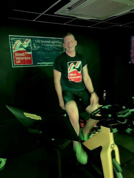 Ian sat on his spin bike wearing his Blind Veterans UK t shirt and with the Blind Veterans UK banner on the wall behind.