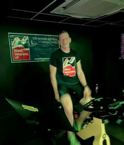 Ian sat on his spin bike wearing his Blind Veterans UK t shirt and with the Blind Veterans UK banner on the wall behind.