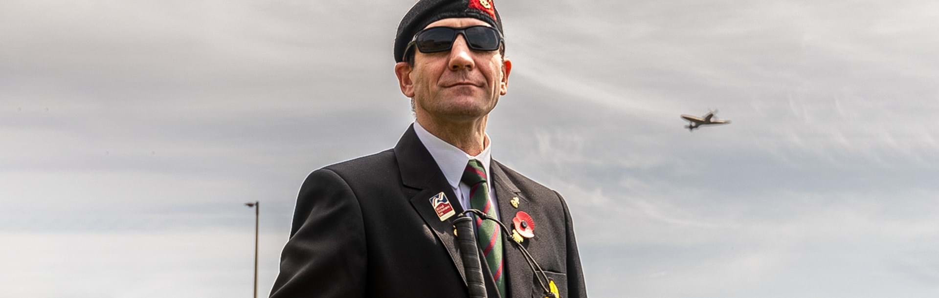 Blind veteran Charlie standing outside wearing a beret, suit and poppy, as a Spitfire flies in the sky above him over our Centre of Wellbeing in Brighton
