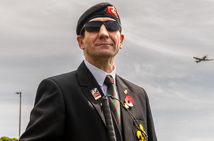 Blind veteran Charlie standing outside wearing a beret, suit and poppy, as a Spitfire flies in the sky above him over our Centre of Wellbeing in Brighton