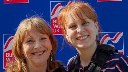 Jennie and Fern are both wearing a harness and standing in front of a Blind Veterans UK branded wall