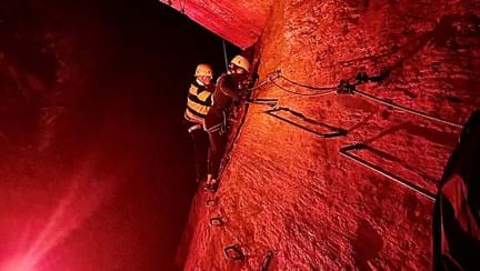 Blind veteran Jules wearing safety gear and a zip-wire harness as he edges along a cavern wall, using footholds and hand barriers