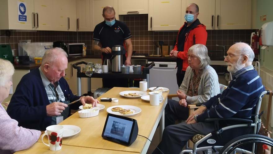 A photo showing a group of blind veterans and their wives enjoying a cup of tea with the scones they baked, assisted by Alexa