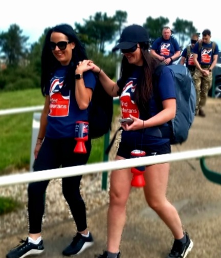 Two women walking along a path. One is wearing a blindfold and the other is guiding her