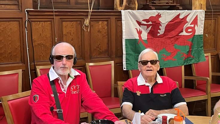 Two men wearing red Wales Rugby shirts sit in front of the Welsh flag which has a white and green background and a red dragon.