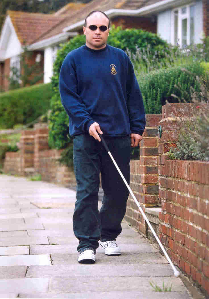 Blind veteran Billy wearing dark sunglasses and using a long white cane as he walks along a path