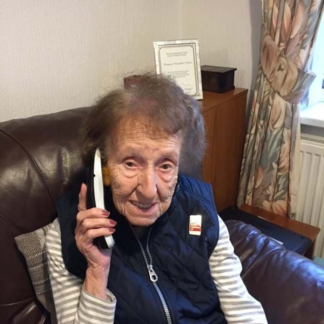 Blind veteran Margaret, sitting on an armchair, smiling to the camera as she holds a phone up to her ear 