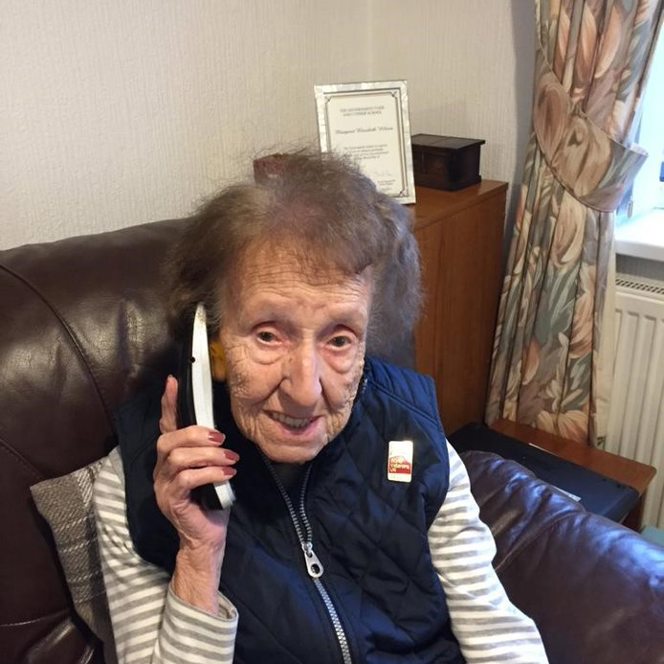Blind veteran Margaret, sitting on an armchair, smiling to the camera as she holds a phone up to her ear