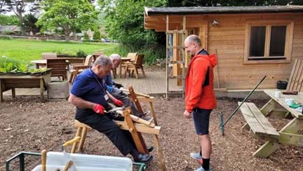 A photo of two blind veterans woodworking, with a wooden cabin in the background