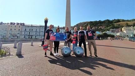 Members of Thea's Surgical Team, wearing Blind Veterans UK branded tops and holding buckets and Blind Veterans UK banners.  They are standing in front of the Llandudno Town War Memorial.  Two giant yoga balls with eyes painted on them can be seen at the front of the group.