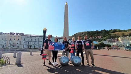 Members of Thea's Surgical Team, wearing Blind Veterans UK branded tops and holding buckets and Blind Veterans UK banners.  They are standing in front of the Llandudno Town War Memorial.  Two giant yoga balls with eyes painted on them can be seen at the front of the group.