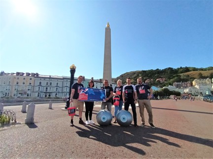 Members of Thea's Surgical Team, wearing Blind Veterans UK branded tops and holding buckets and Blind Veterans UK banners. They are standing in front of the Llandudno Town War Memorial. Two giant yoga balls with eyes painted on them can be seen at the front of the group.