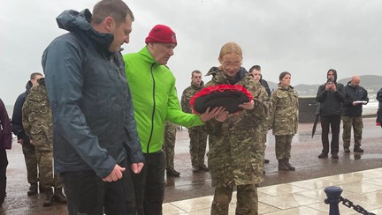 Blind veteran John wearing a bright yellow jacket lays a wreath with a charity volunteer alongside him and a student from the Military Preparation College of training on the other side.