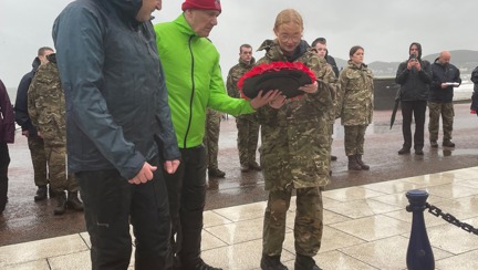 Blind veteran John wearing a bright yellow jacket lays a wreath with a charity volunteer alongside him and a student from the Military Preparation College of training on the other side.