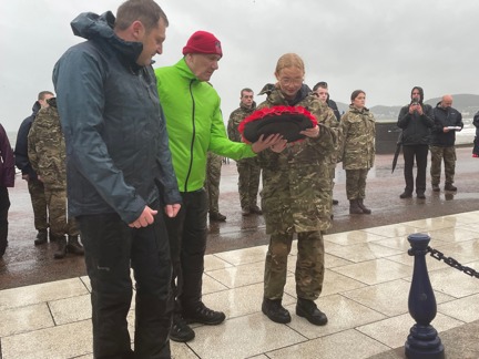 Blind veteran John wearing a bright yellow jacket lays a wreath with a charity volunteer alongside him and a student from the Military Preparation College of training on the other side.