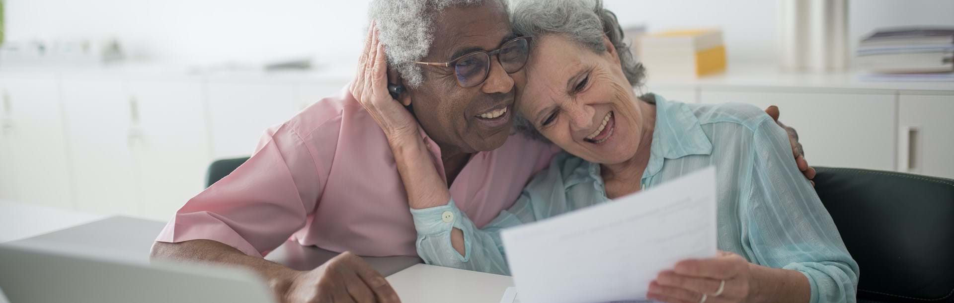 An elderly couple laughing while holding a document and using a laptop