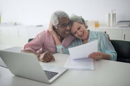 A photo of an elderly couple laughing while holding a document and using a laptop