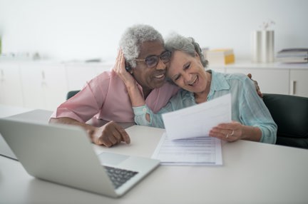 An elderly couple laughing while holding a document and using a laptop