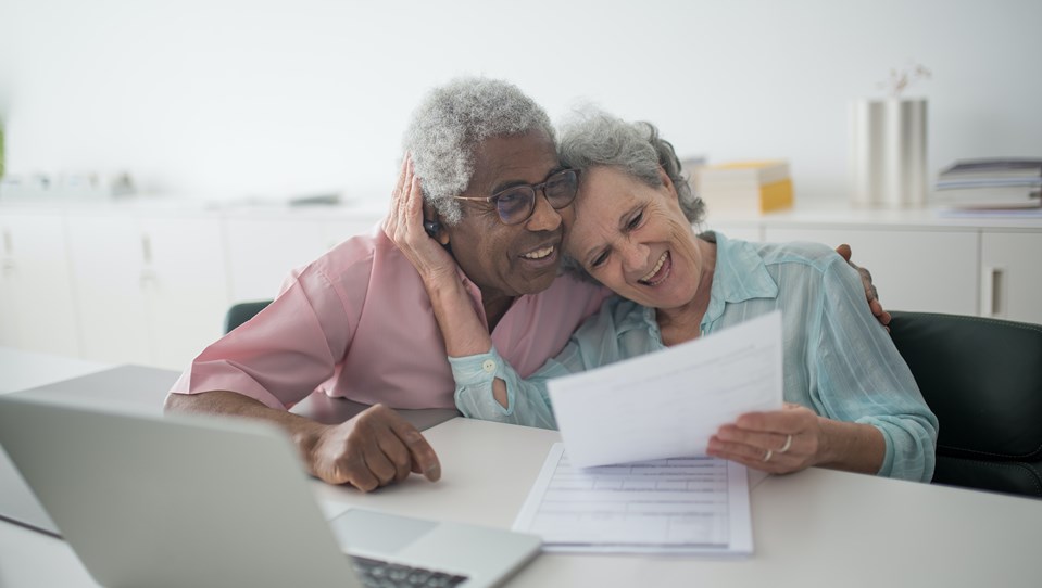 An elderly couple laughing while holding a document and using a laptop