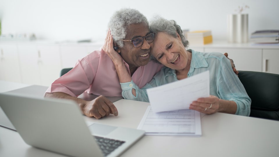 An elderly couple laughing while holding a document and using a laptop