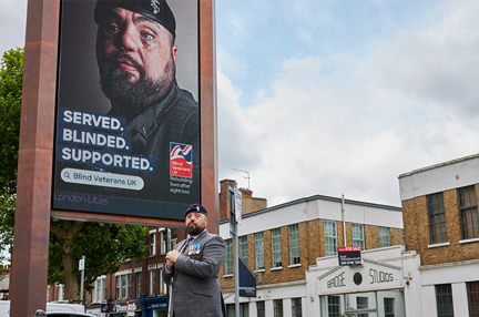 Blind veteran Simon is wearing a blazer, beret and medals and stands side on in front of a large digital screen which has an advert of himself and the line Served, Blinded, Supported