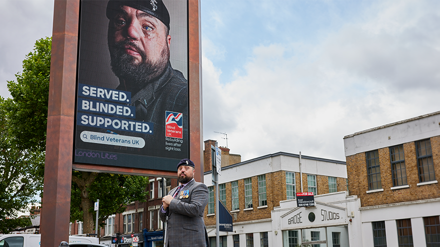 Blind veteran Simon is wearing a blazer, beret and medals and stands side on in front of a large digital screen which has an advert of himself and the line Served, Blinded, Supported