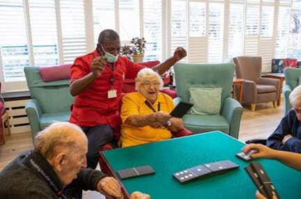 Blind veteran Win smiling, sat a table with other veterans with a support worker resting on the arm of her chair, cheering, as she places a giant domino on the table.