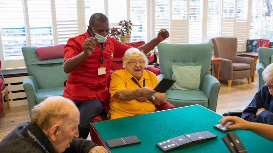 Blind veteran Win smiling, sat a table with other veterans with a support worker resting on the arm of her chair, cheering, as she places a giant domino on the table.